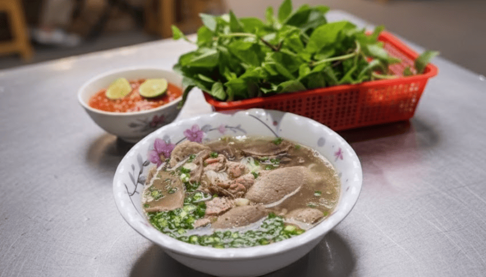 A bowl of Vietnamese beef pho with sliced beef and scallions next to fresh green herbs and red chili dip with lime slices.