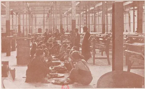 Historical sepia photograph of workers sitting on the floor having a meal inside a large industrial factory hall.