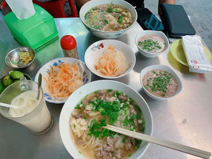 Bowls of Vietnamese beef noodle soup served on a metal table with sliced pickled onions, green herbs, dipping sauces, and an iced drink.