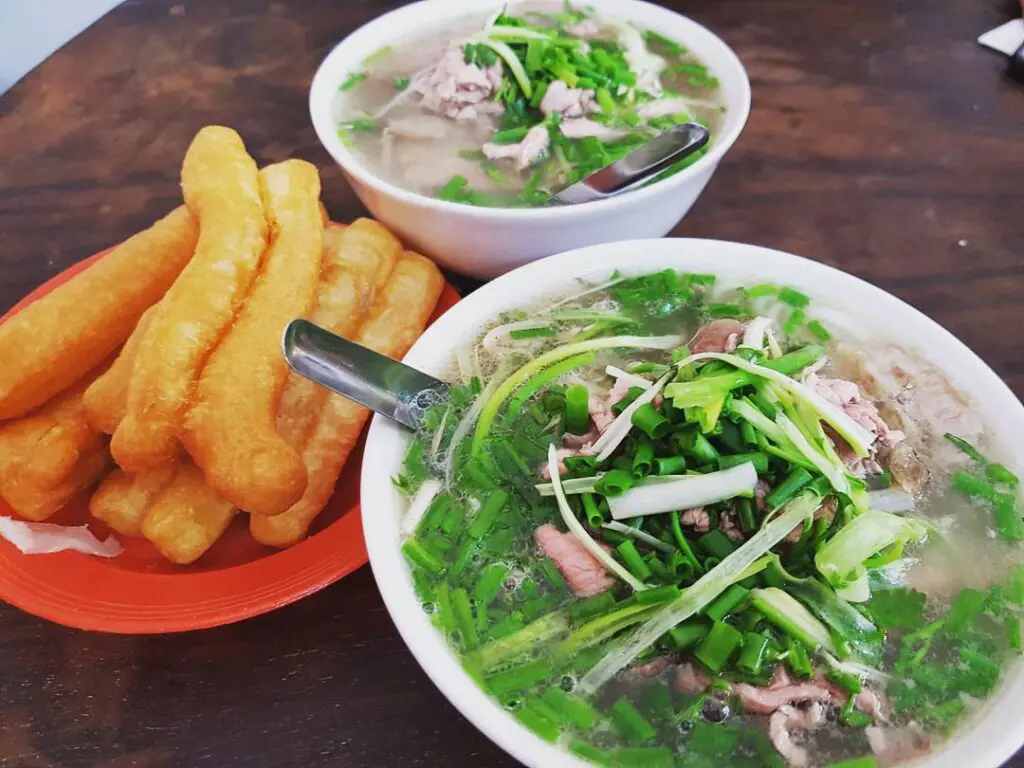 Two white bowls of hot beef pho noodle soup topped with green scallions, served with golden fried dough sticks on a red plate.
