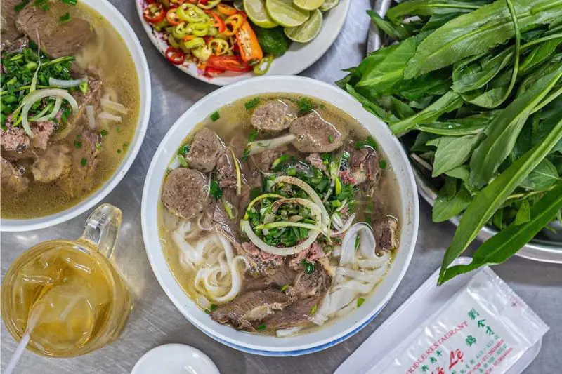 Top-down view of bowls of Vietnamese beef noodle soup served with beef meatballs, fresh herbs, lime, and iced tea.