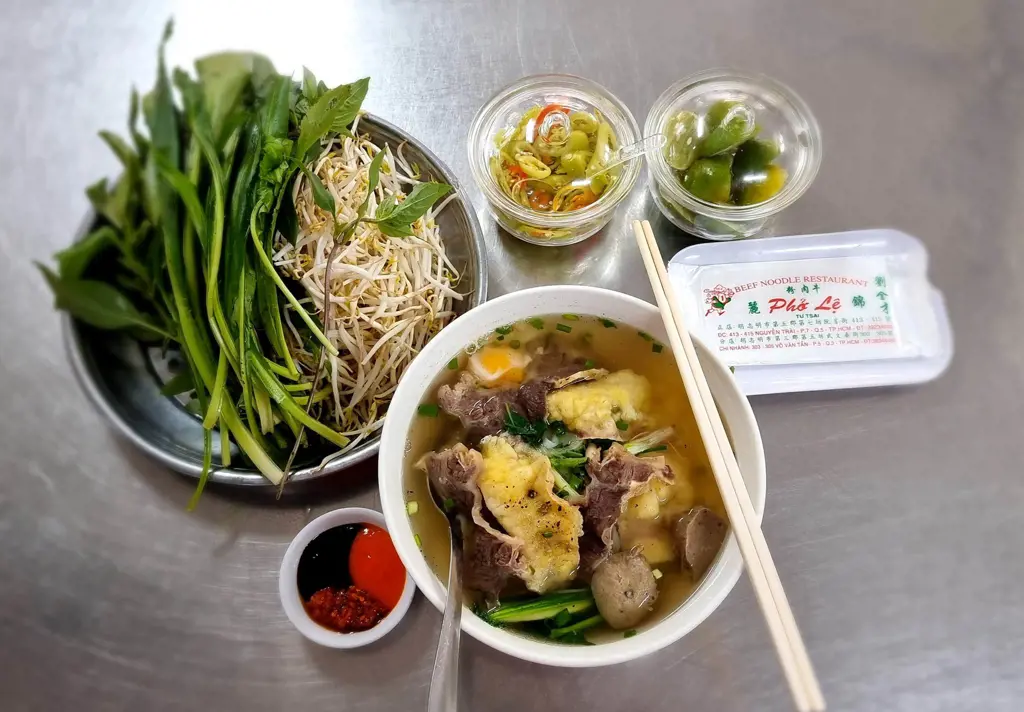A bowl of Vietnamese beef noodle soup with beef slices, meatballs, and an egg yolk, served with fresh green herbs, bean sprouts, and condiments.