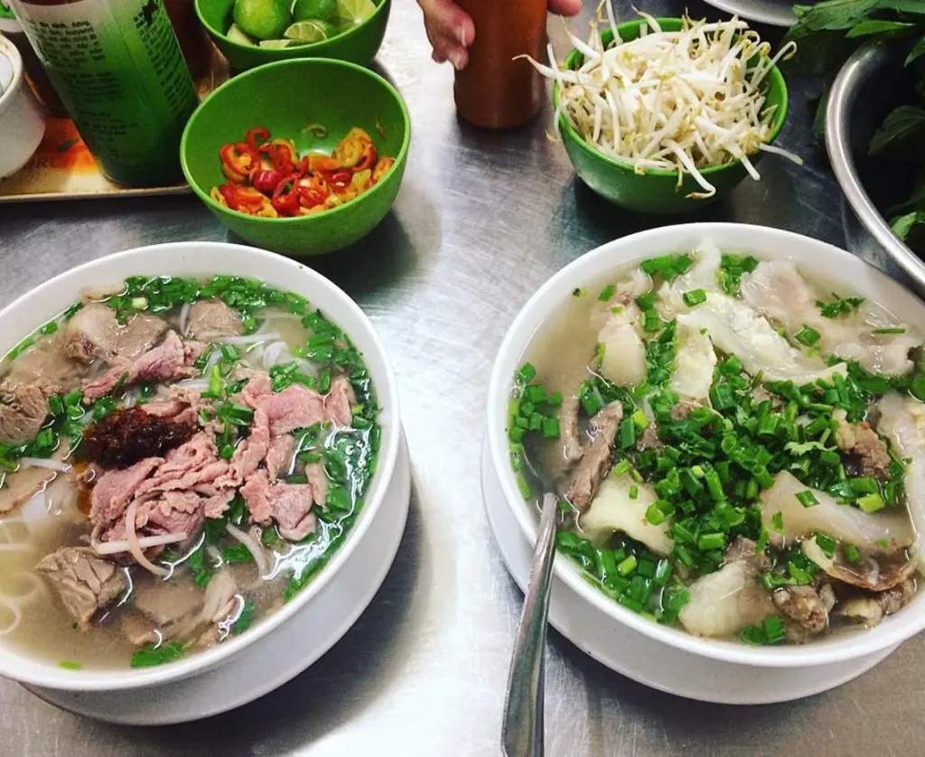 Two bowls of Vietnamese beef pho noodle soup served with chili slices, bean sprouts, and lime on a metal table.