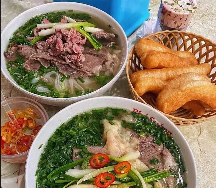 Two bowls of hot Vietnamese beef pho garnished with abundant scallions and sliced chillies, served with golden fried dough sticks in a small basket.