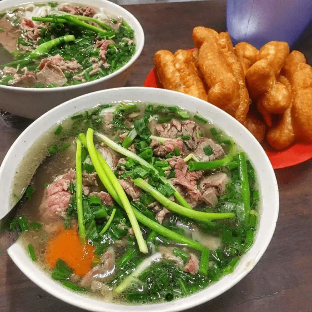 Two bowls of Vietnamese beef pho topped with green scallions and sliced beef, accompanied by a plate of golden fried dough sticks.