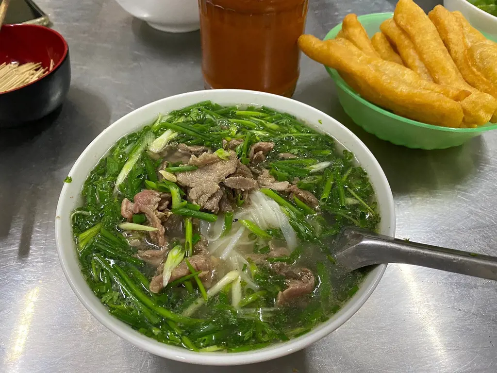 A bowl of Vietnamese beef pho overflowing with chopped scallions and noodles on a metal table next to fried dough sticks.