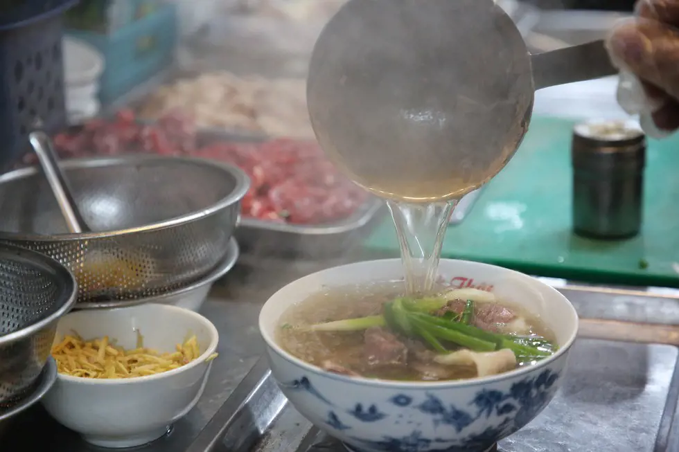 A metal ladle pouring hot broth into a bowl of pho with beef and green onions.