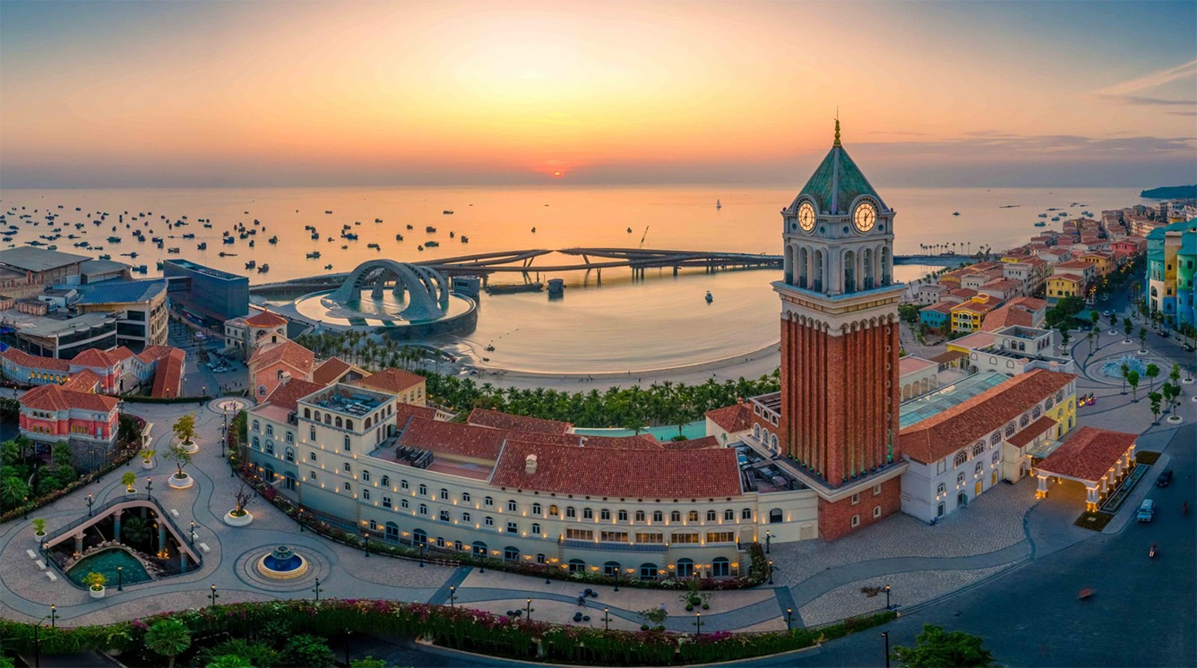 Aerial view of Sunset Town in Phu Quoc during golden hour, showcasing a tall clock tower, terracotta-roofed coastal buildings, Kiss Bridge, and boats on the sea.