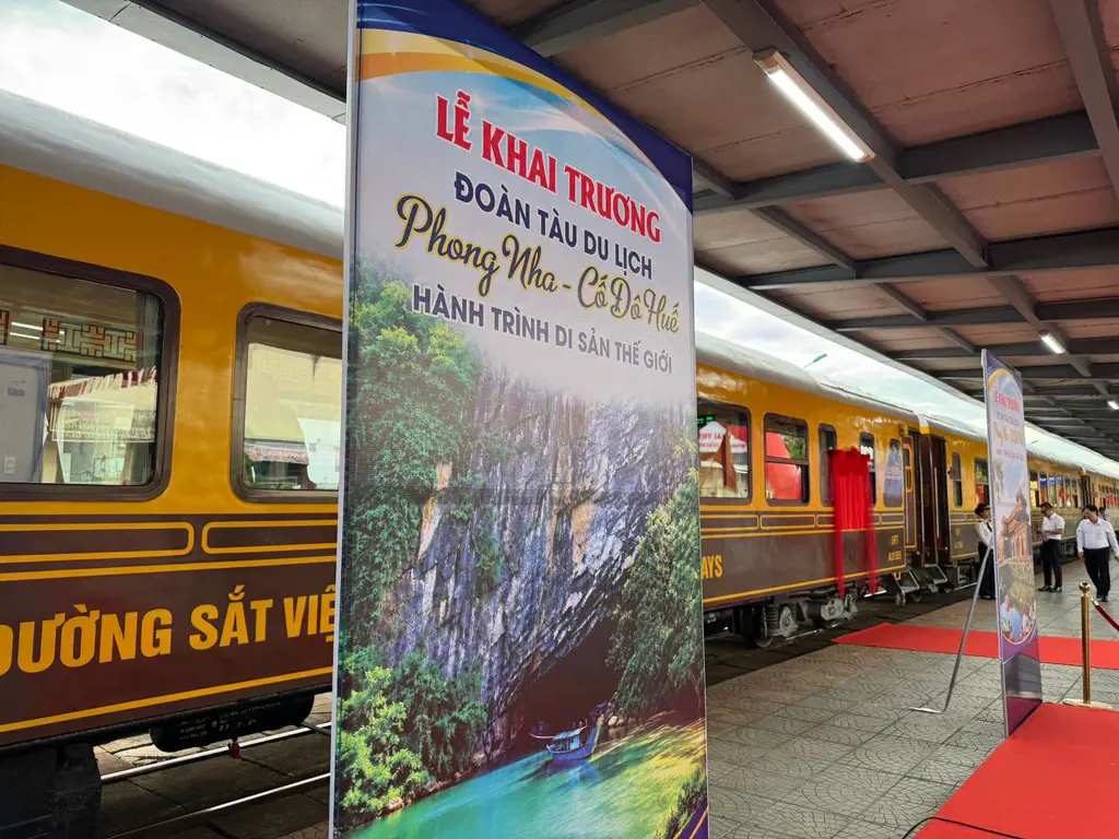 Tấm banner quảng bá lễ khai trương đoàn tàu du lịch Phong Nha - Cố Đô Huế đặt trên sân ga bên cạnh toa tàu màu vàng và nâu.