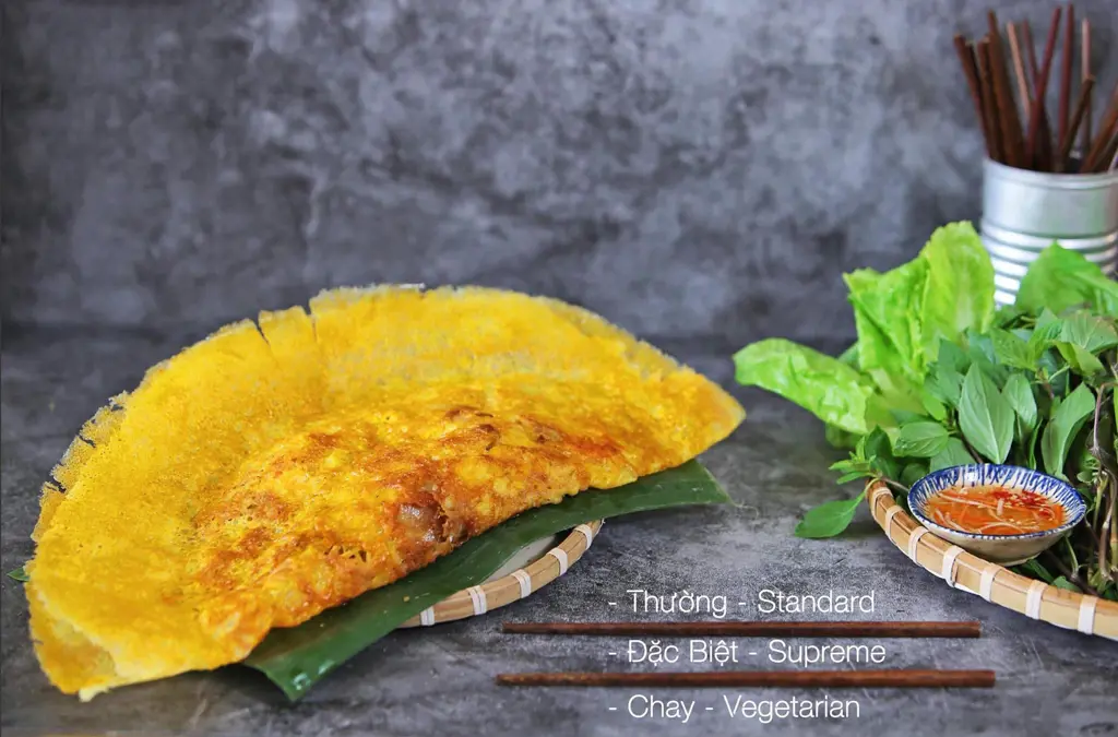 A golden crispy Vietnamese Bánh Xèo pancake served on a banana leaf next to a basket of fresh herbs, lettuce, dipping sauce, and chopsticks.