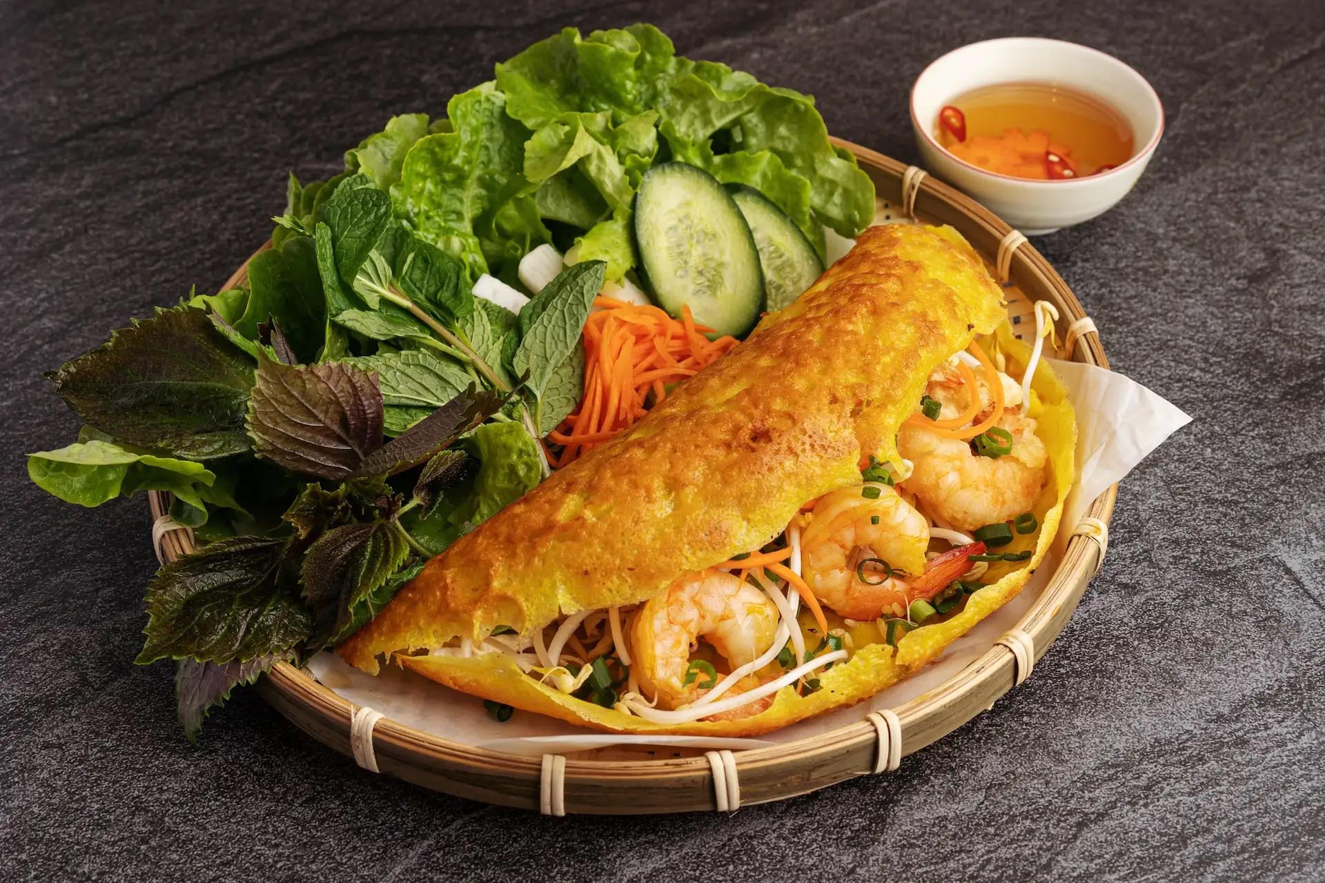 A golden crispy Vietnamese pancake filled with cooked shrimp and bean sprouts, presented on a bamboo platter with fresh greens and dipping sauce.