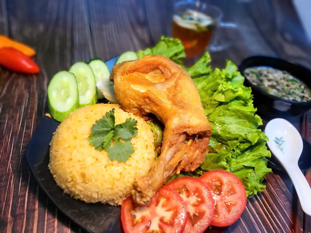 A plate of crispy fried chicken leg paired with a dome of yellow rice, tomato slices, cucumber slices, and lettuce.