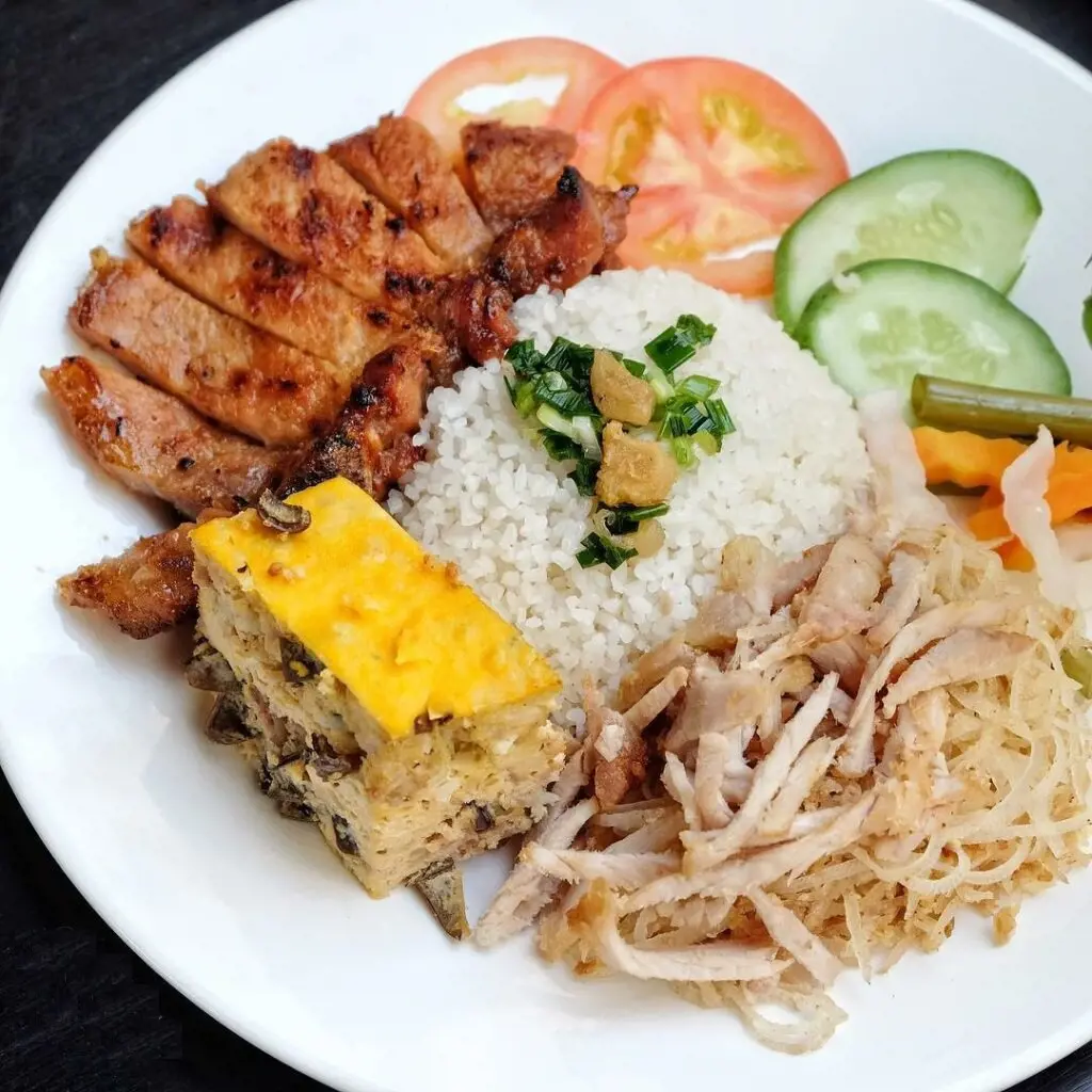 A white plate featuring Vietnamese broken rice topped with scallion oil, accompanied by sliced grilled pork chop, steamed egg meatloaf, shredded pork skin, tomato slices, and cucumber slices.