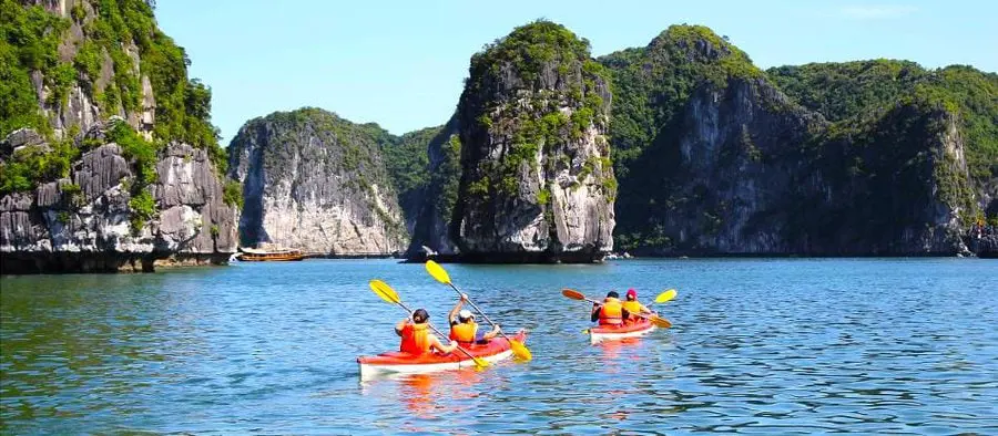 Tourists paddling red kayaks in clear blue waters among towering green limestone karst mountains in Lan Ha Bay.