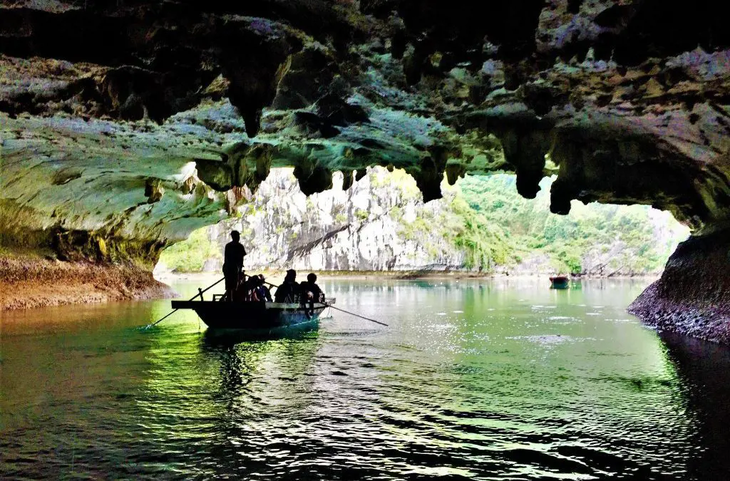 A boat carrying passengers glides under stalactites inside Dark Cave in Lan Ha Bay, looking out toward emerald water and limestone mountains.
